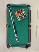 Mini pool table with green felt, six netted pockets, surrounded by dark wood frame, with a cue stick and full billiard ball set arranged on the playing field.