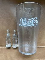 Photo shows one Pepsi Cola plastic cup with textured surface and white logo along with two small clear glass Coca-Cola bottles without labels or caps, all placed on tan leather surface.