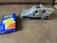 Polaroid Joy Cam camera with Polaroid 500 Joycam film box placed on a tan surface.