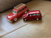 Photo of two red Volkswagen toy cars on a couch surface. Left is a VW Type 3 Squareback station wagon toy car in red with roof rack. Right is a smaller VW Microbus toy car in red.