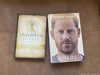 Two books placed side by side on a brown couch: 'Shattered Silence' on the left and 'Spare' on the right.