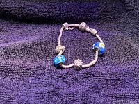 Pandora sterling silver charm bracelet on dark fabric background, showing six diverse charms including silver and blue glass bead charms.