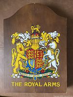 Photo of the closed wooden dart board cabinet showing the painted crest 'The Royal Arms' with lion and unicorn figures, crown, and motto.