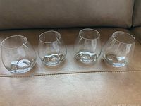 Photo of four clear stemless wine glasses arranged in a row on a fabric surface, showing their rounded bowl shapes and smooth rims.