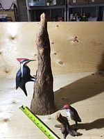 Front view of two bird carvings, large Pileated Woodpecker on textured tree trunk, smaller bird on wood base, measuring tape for scale.