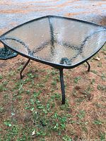 Photo showing the square textured glass top patio table with metal frame and four legs on outdoor ground with pine needles and dirt.