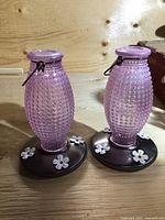 Two pink pebbled glass hummingbird feeders with black bases and white flower decorations shown side by side on a wood surface.