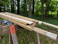 Assorted new lumber of various widths resting on two wooden saw horses with orange metal brackets in an outdoor grassy area.