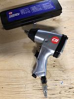 Silver and black 1/2 inch Campbell-Hausfeld air impact wrench with logo, placed on wooden surface beside closed black metal case with blue label.