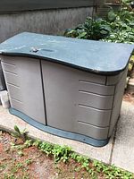 Front and side view of outdoor storage box showing green lid and beige body with horizontal groove details, placed on a concrete patio.