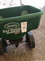 Side view of green hopper with Scotts brand logo and Turf Builder EdgeGuard DLX label, showing the large wheel beneath.
