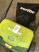 Top view of SunJoe green electric lawn aerator with attached collection bag, showing brand name and unit body condition.