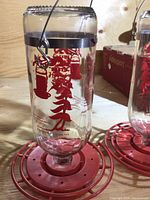 Two large glass hummingbird feeders with clear glass bodies, red graphic decoration, red plastic circular bases with multiple feeding ports and perches, metal hanging hooks, and clear plastic ant moats with hanging hooks.