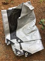 Close-up of a folded silver/gray tarp showing black reinforced corners with white metal grommets, on outdoor ground surface.