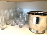 Various clear glass barware including tall highball glasses, short rocks glasses, square and round glass styles with a hammered metal chill bucket with handles.