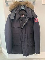Front view of jacket on hanger showing fur-trimmed hood, flap pockets, button and zip closure, Canada Goose sleeve logo patch.