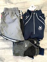 Photo of folded grey Lululemon shorts, navy blue Yonex sports jacket, and grey North Face sleeveless top on wooden floor.