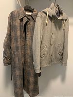 Front view of plaid trench coat on hanger and gray hooded collegiate jacket on hanger side by side