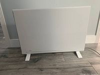 Front view of a white slim style Delonghi convection panel heater standing on two feet on a wooden floor.