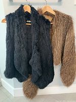 Two fur vests hanging on wooden hangers against a white wall, black Alice + Olivia vest in front and brown Michael Kors vest behind.