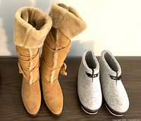 Both pairs of ladies boots displayed side by side on a dark wooden surface under soft light: tan Tod's boots with fur collar and gray Scandinavian booties with buckle detail.