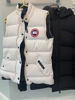 White Canada Goose vest and black jacket hanging side by side, front view