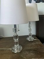 Pair of crystal base lamps with white drum shades on wooden surface.