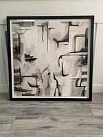 Full view of large framed abstract print on floor with black frame and neutral abstract design in black, gray, and brown tones.
