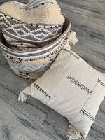 Photo showing one fabric nesting basket with beige and brown geometric patterns and sequins with the beige decorative cushion with tassels on the floor.