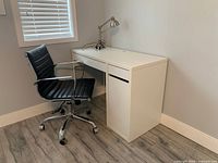 White office desk with black swivel chair and chrome desk lamp placed beside it in a room with hardwood flooring.
