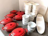 Seven red ramikin mini casseroles with lids, nine white ramekins, one red mini Crock Pot, and two long white serving plates on glass table.