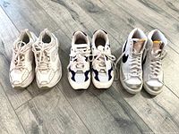 Photo of three pairs of sneakers: white sneakers, New Balance white/navy sneakers, and white Nike high tops on a wooden floor.