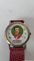 Close-up view of watch face featuring Betty Boop illustration with green background and black numerals on white dial, silver-tone bezel, and red textured strap.