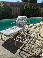 Two 1950's Mid Century aluminum chaise lounges shown poolside. One chaise lounge with floral cushion and one without cushion showing frame and webbing.