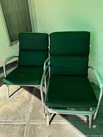 Two 1950s aluminum glider lounge chairs with green vinyl cushions placed side by side on a tiled floor outdoors.