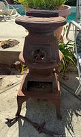 Side view of antique cast iron potbelly stove showing front vent door and legs, with one leg detached and laying on ground.