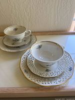 Two vintage Schumann Germany teacup sets each showing a teacup, a saucer, and a decorative pierced plate with blue floral patterns and gold trim on a wooden surface against a textured wall.