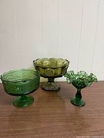 Three olive green glass pieces: one compote and two candy dishes, shown on a wooden surface against a white wall.