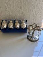 Four silver-tone salt and pepper shakers in blue box next to metal hanger with two hanging glass grape-shaped shakers.