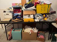 Large assortment of folded vintage textiles, fabric rolls, and a yellow plastic tub filled with cloths stacked on a black table. Lower shelf holds cardboard boxes with additional textiles and sewing items.