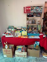 Full view of table covered with red cloth showing large assortment of books, toys, board games, and art supplies along with cardboard boxes filled with more items beneath and next to table.
