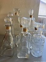 Lot of six clear glass vintage decanter liquor bottles with cork stoppers seen from angled side view on table, some showing intricate pressed glass patterns and different stopper shapes.