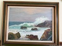 Framed oil painting showing ocean waves crashing against rocks with artist signature visible.