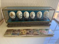Front view of the glass case holding six painted Chinese decorative eggs and seven painted ceramic tiles below.