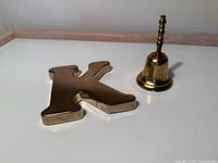 Photo showing a brass hand bell with the engraving "John F. Kennedy" and a brass letter K paperweight on a white surface.