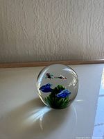 Front angled view of the clear glass paperweight showing blue flowers, green leaves, and two butterflies inside against a light background.