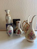Front view of five porcelain pieces showing varied floral designs and shapes including the ribbed sake bottle and small vases with boxed item in background.