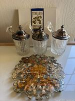 Three ribbed glass jam jars with silver-tone metal lids and teaspoons inside, displayed with ornate silverplate serving pieces