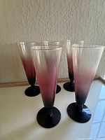 Five vintage champagne flute glasses with ombre frosted pink to dark plum color, standing on a flat surface against a textured wall and window light.