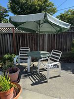 Photo of a vintage patio set outdoors, featuring two white chairs with horizontal slats, a round glass-top table with a green frame, and a large green umbrella.